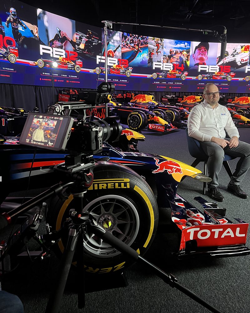 The B-camera on sticks beside a Red Bull Racing car, with a spokesperson seated beyond it